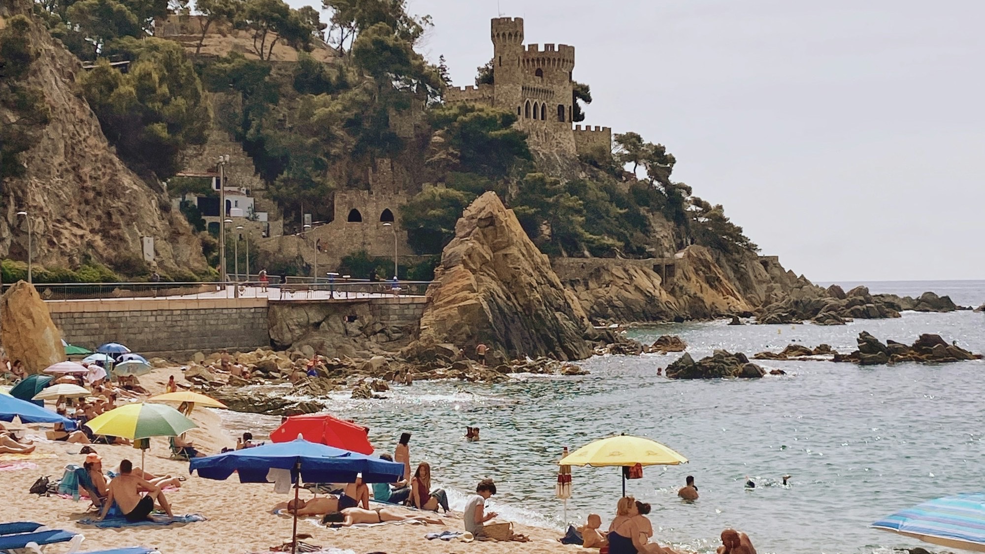 Cala de Sa Caleta en Lloret de Mar con el Castell d'en Plaja al fondo, sombrillas de colores en la arena y bañistas en el mar